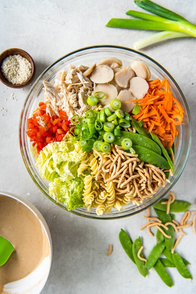 A large glass bowl filled with chow mein noodles, carrots, bell pepper, cabbage, pasta, chicken and water chestnuts. 