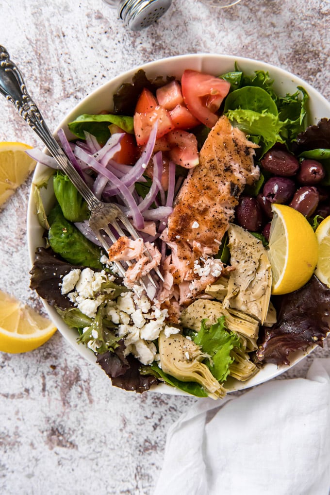 An overhead image of a bowl of grilled salmon salad with olives, tomatoes, feta and artichokes and lemon