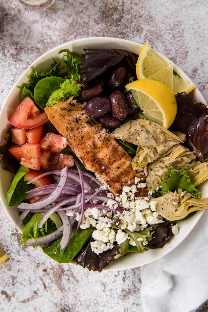 An overhead image of a bowl with lettuce, salmon, artichokes, tomatoes, onions and feta cheese