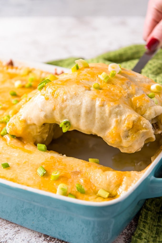 A spatula lifting two enchiladas out of a full pan. 