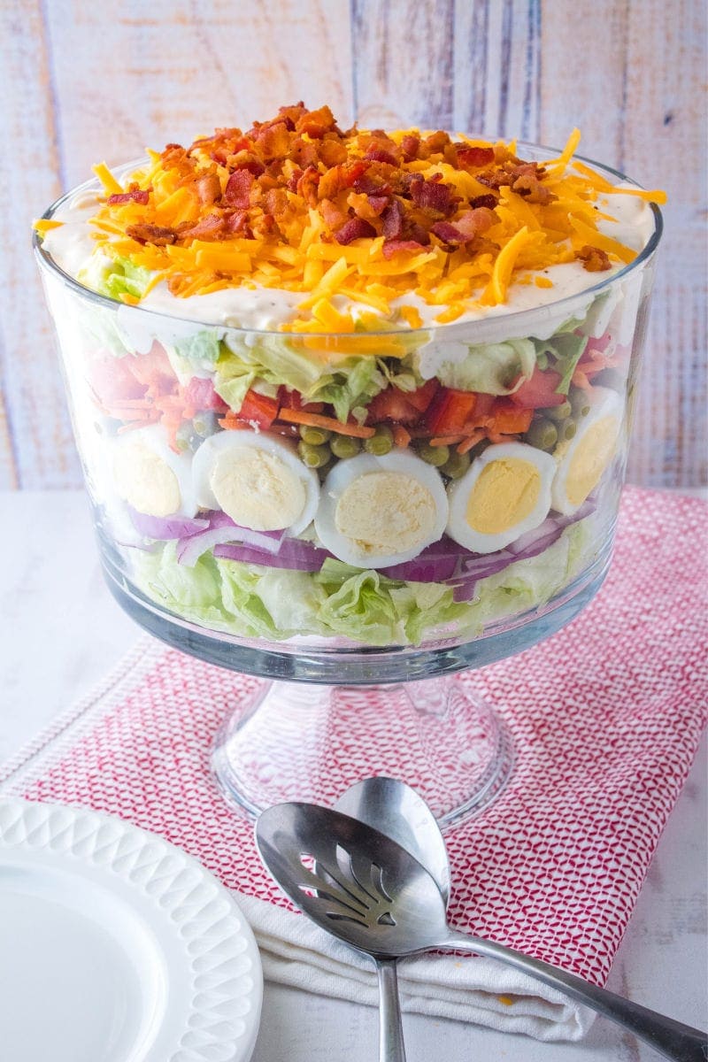 7 Layer Salad is a classic potluck dish great for feeding a crowd. These layers of deliciousness can be made ahead of time for these easiest side dish ever! layer salad in a trifle bowl with serving spoons and plates