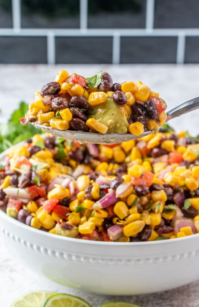 large bowl of black beans, corn, tomatoes and avocado salad with a metal spoon lifting up a serving.