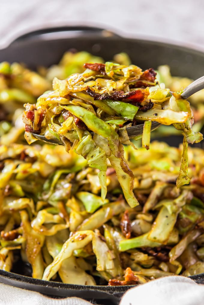 Shredded cabbage and bacon, fried together in a cast iron skillet. A serving is held up by a metal spoon.