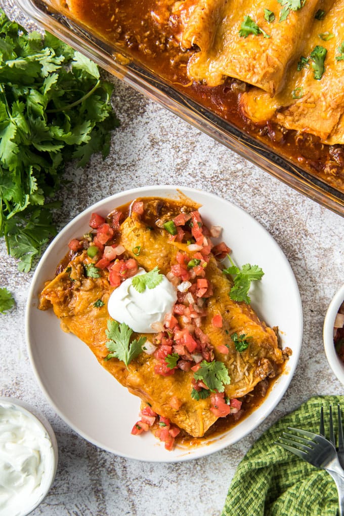 an overhead image of two enchiladas on a plate next to a large dish of enchiladas