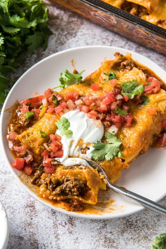 Two beef enchiladas on a plate with a fork taking a pieces out of one of them