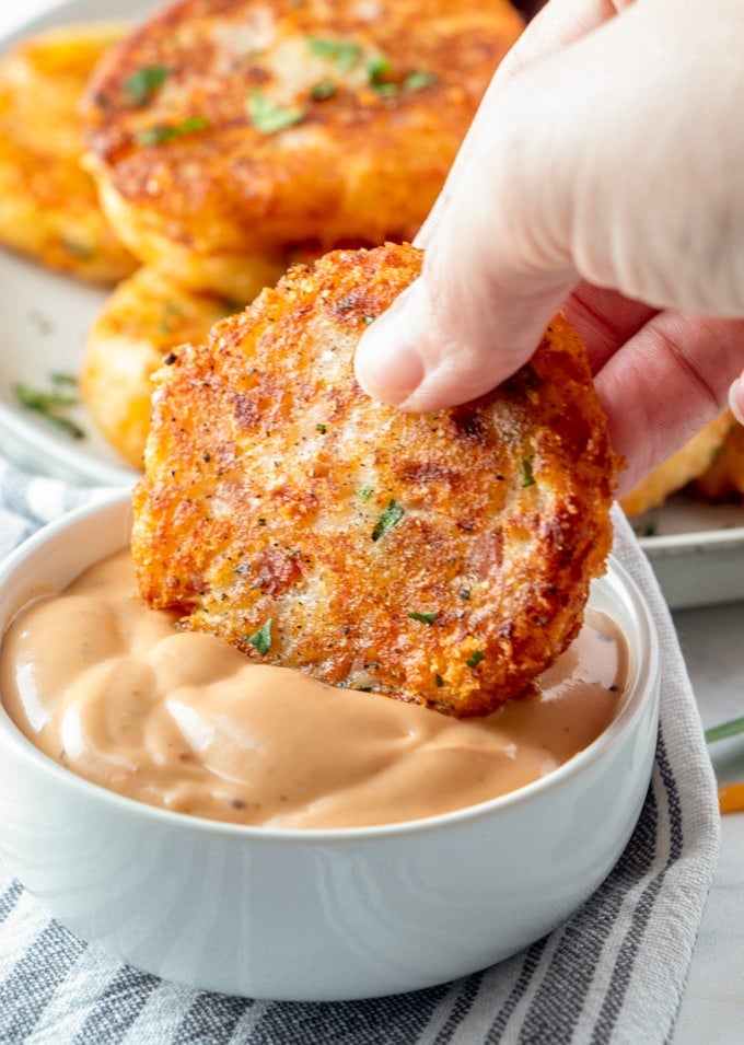 a hand holding a potato croquette being dipped into an orange sauce