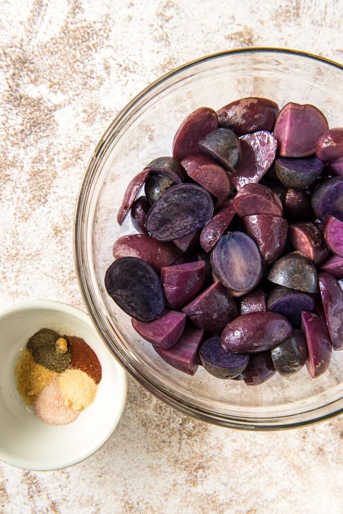 a clear glass bowl filled with cut up purple potatoes and a small white dish with powdered seasonings in it.
