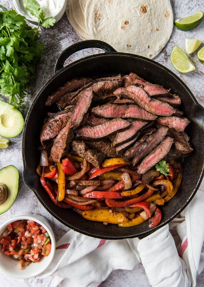 un'inquadratura dall'alto della bistecca affettata e dei peperoni in una padella, circondata da salsa, avocado e tortillas