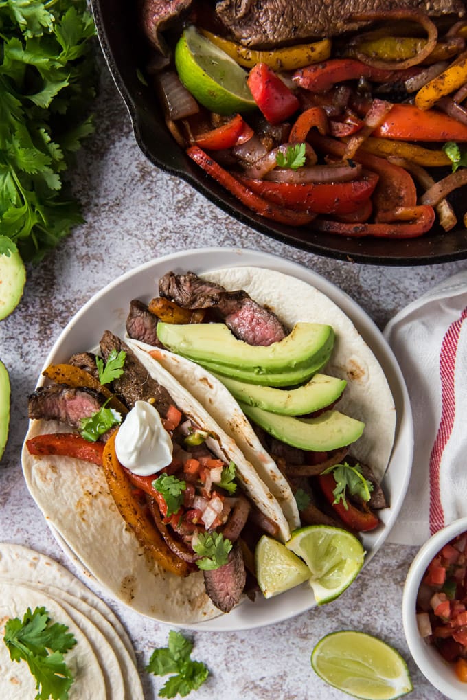 Une assiette blanche avec deux tortillas de farine remplies de steak et de légumes, garnies de crème sure et de salsa. Une poêle à côté contient du steak et des poivrons.