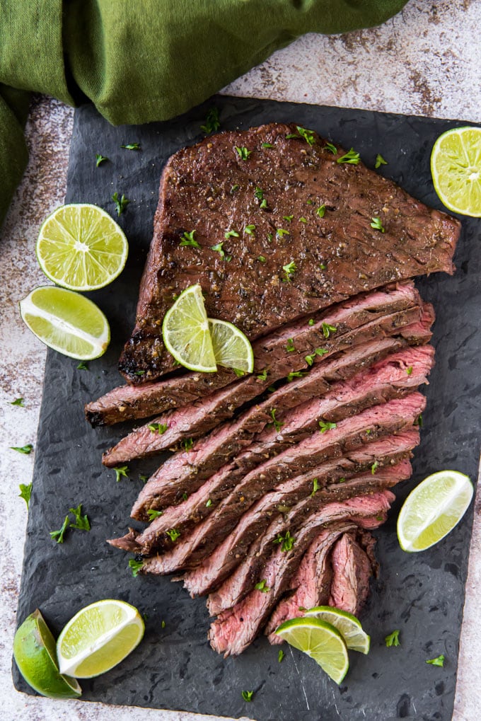 an overhead image of a large marinated and grilled flank steak, cut into strips and surrounded by sliced limes