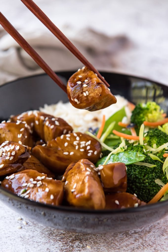 chopsticks holding a piece of teriyaki chicken over a bowl of rice and veggies