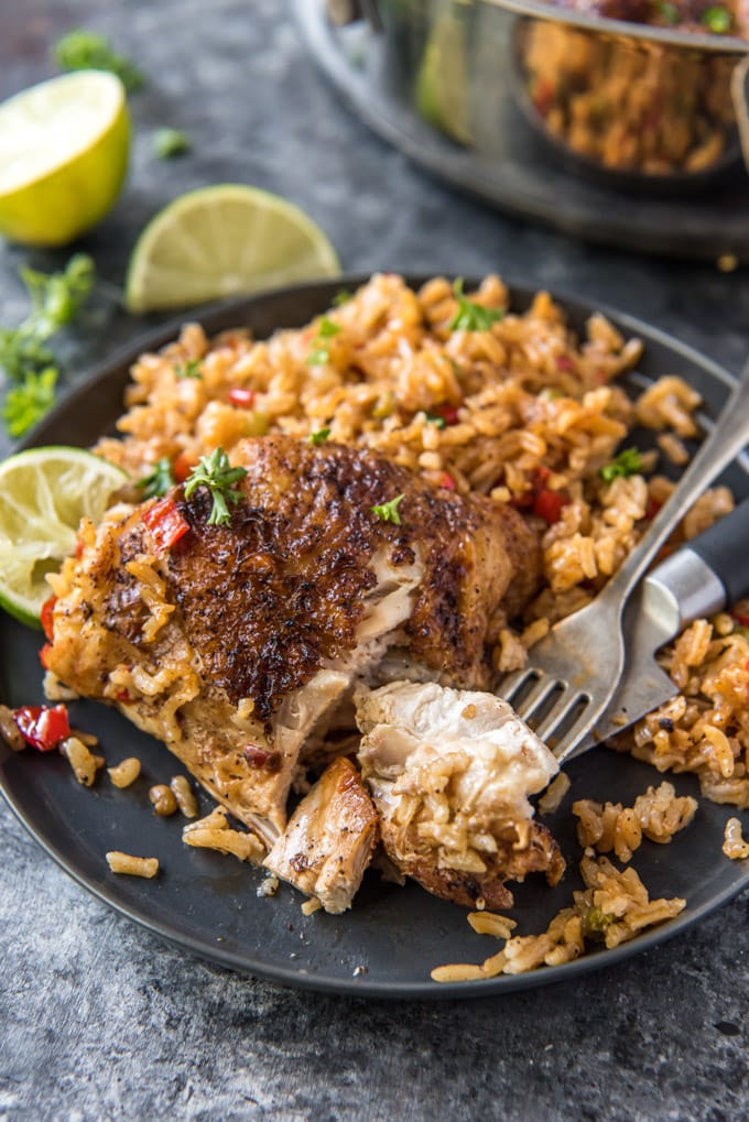 A chicken thigh on a black plate next to a bed of rice with a piece cut off and sitting on a fork.