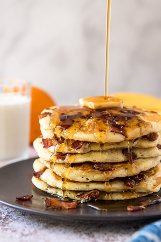 A stack of bacon pancakes with syrup being poured over the top.
