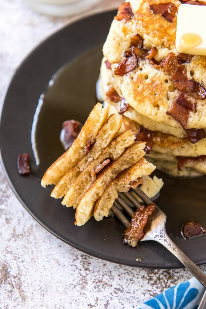 A fork laying on a black plate with pieces of a cut up pancake with bacon