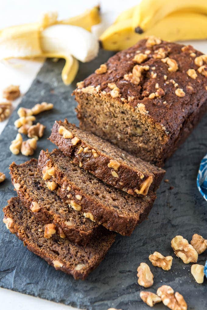 Rebanadas de pan de plátano y nueces cortadas de un pan con plátano en el fondo.