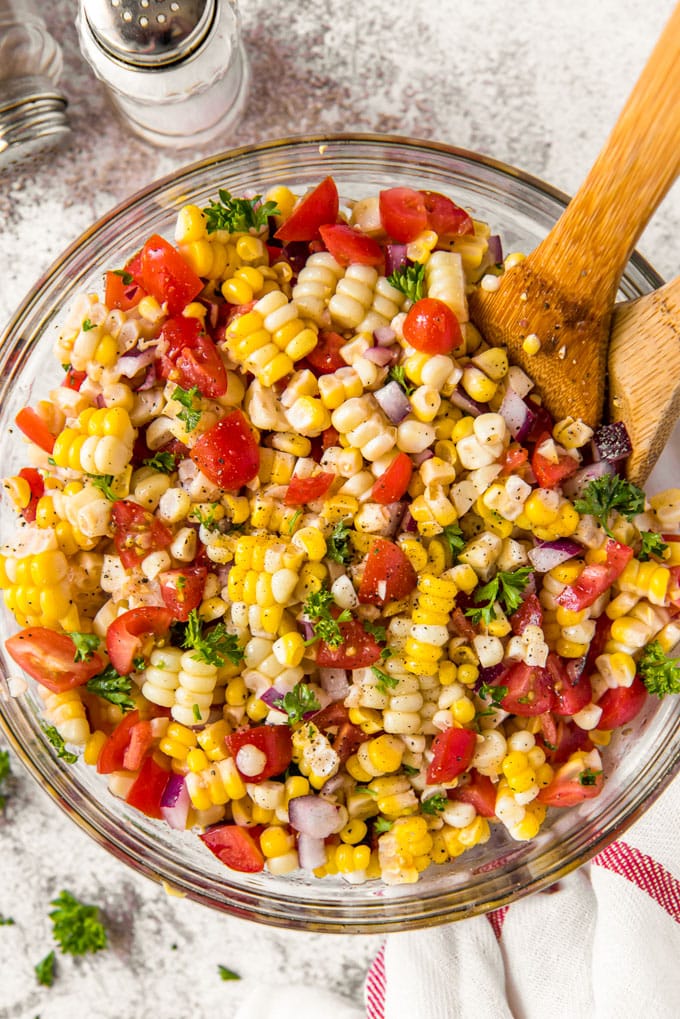 A bowl of corn salad with tomatoes, parsley and onions with two wood spoons sticking out.