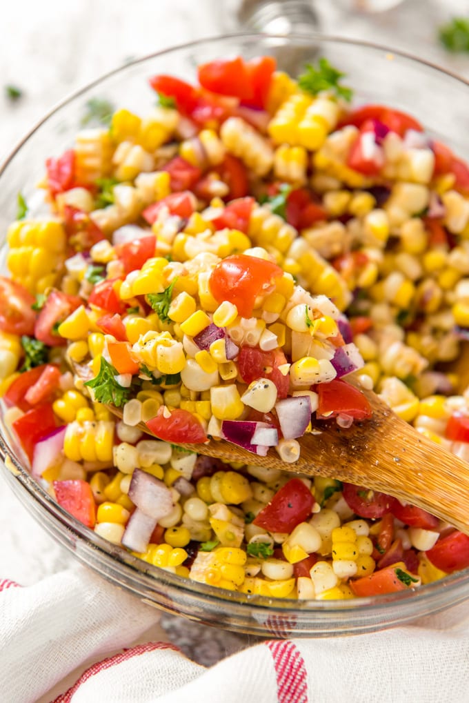A large glass bowl filled with chopped corn, tomatoes and onions and a wooden spoon.
