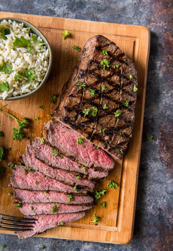 A large cutting board featuring a whole London Broil grilled steak, sliced, with a small bowl of rice.