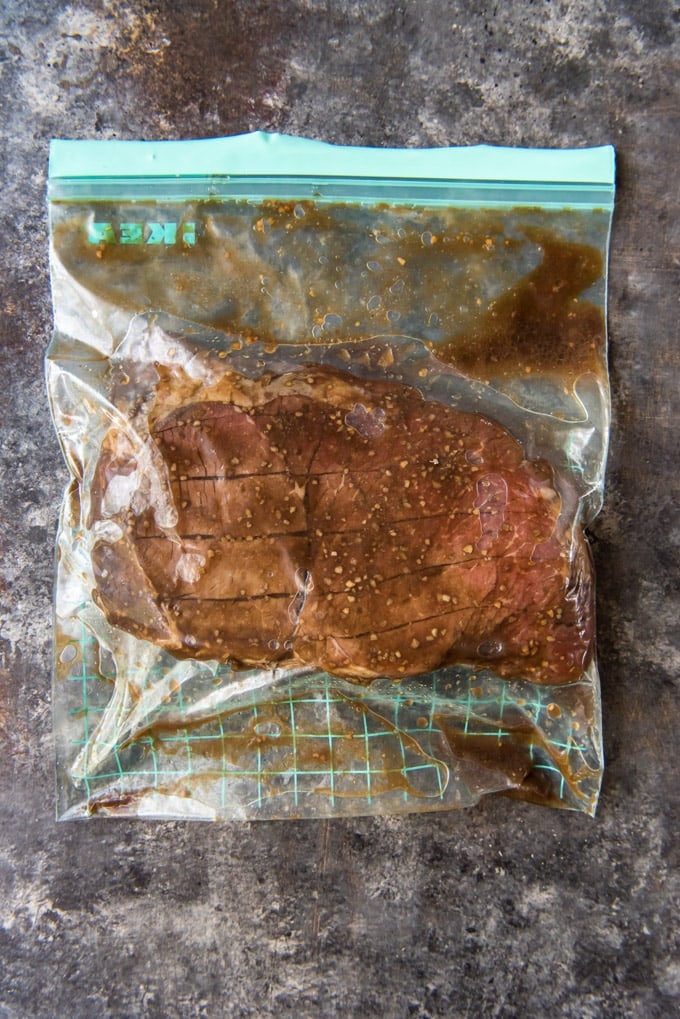 A large steak that has slits cut into it, sitting in a marinade in a resealable plastic bag.