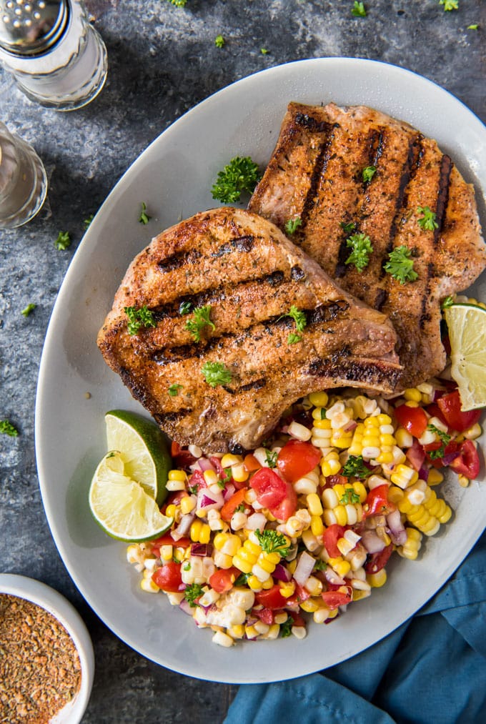 a large white plate with two grilled pork chops and corn salad on the side.