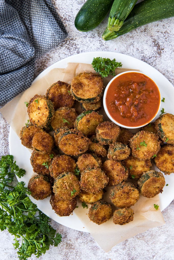 An overhead image of a white plate with a pile of fried zucchini slices and a bowl of marinara sauce. A blue and white napkin is off to the side, across from 3 whole zucchini. 