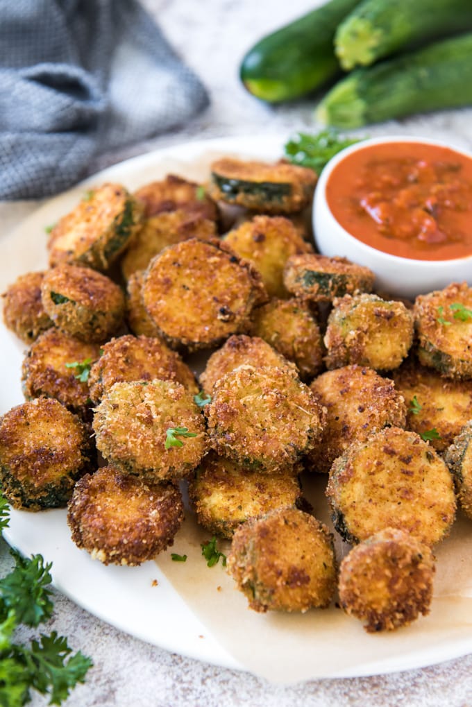 a white plate with a pile of fried zucchini and a small bowl of marinara. A blue and white napkin and some whole zucchini are in the background.