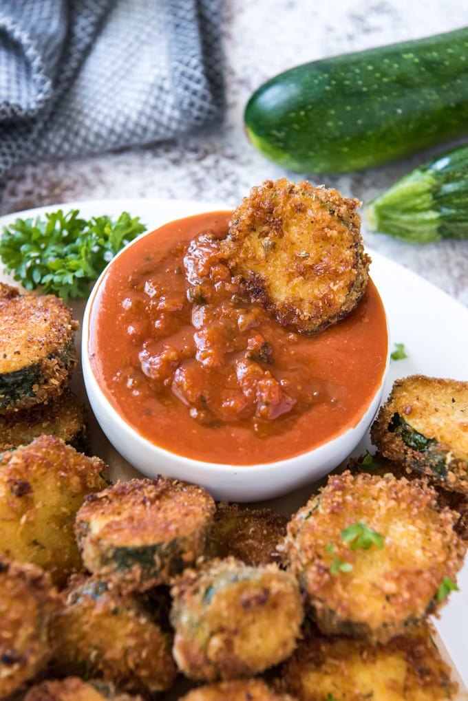 a small bowl of marianara with a piece of fried zucchini dipping in. surround by more fried zucchini slices on a plate.