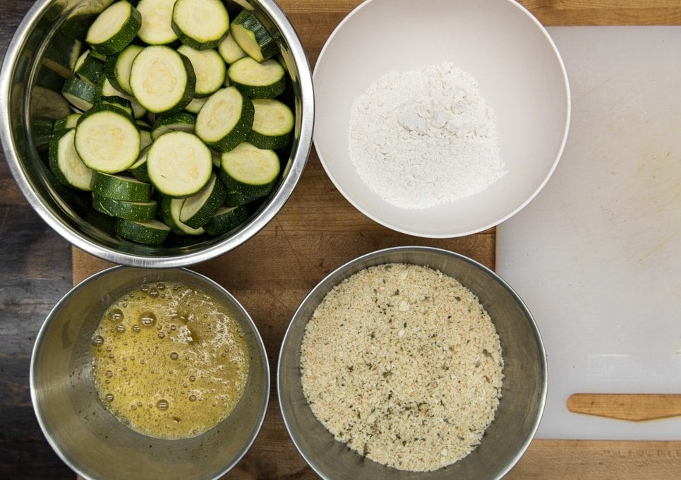 4 bowls with sliced zucchini, flour, whisk egg, and breadcrumbs.