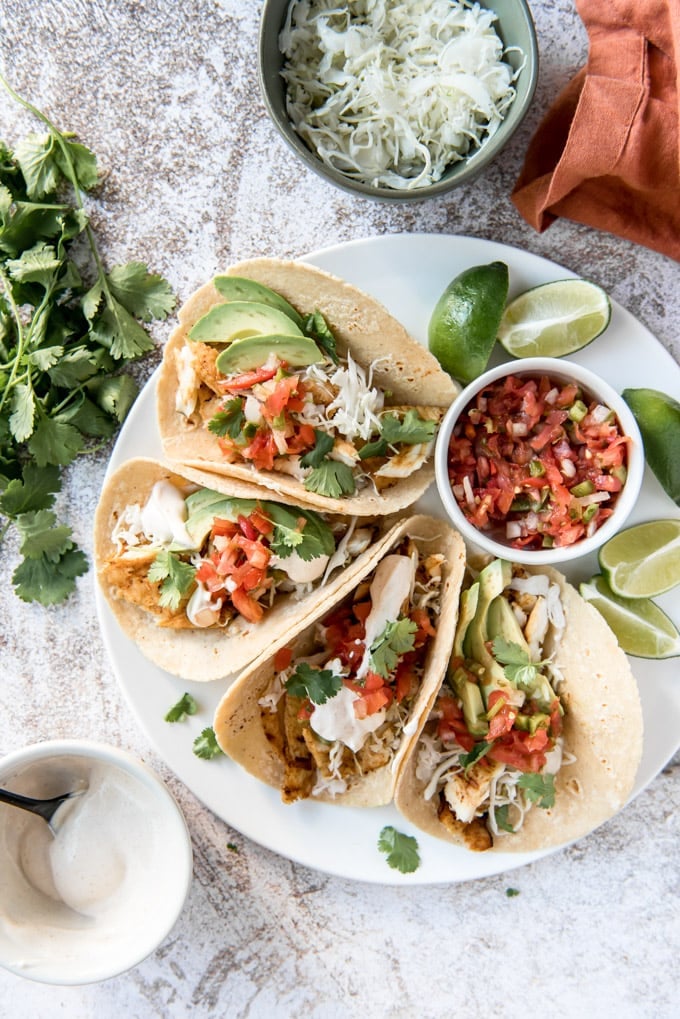 An overhead image of 4 fish tacos on a white plate with sliced limes, with a bowl of sauce, a bowl of cabbage and a napkin in the background.