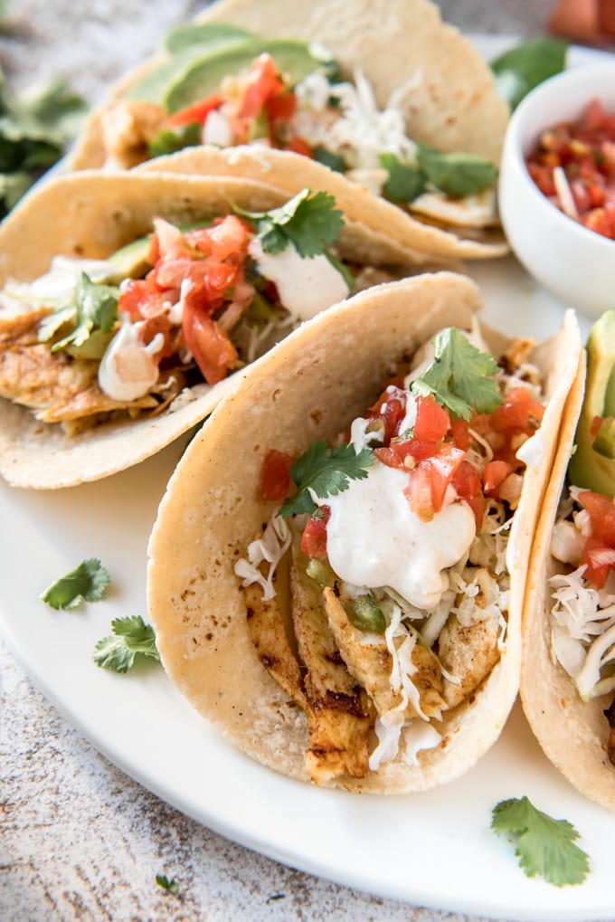 a white plate with corn tortillas filled with grilled fish and topped with cilantro, sour cream and tomatoes.