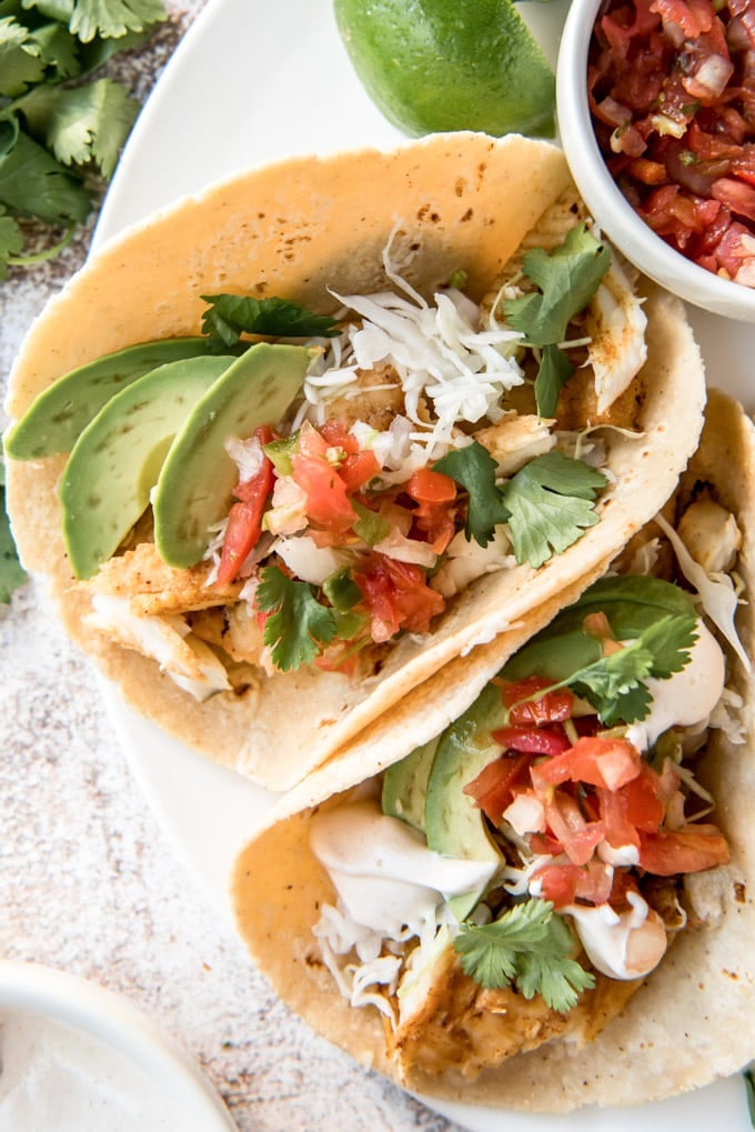 A close up overhead image of grilled fish tacos, stuffed with avocado slices, cilantro and pico de gallo on corn tortillas.