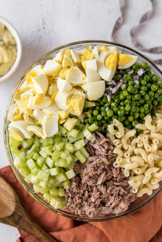 a large glass bowl with ingredients for tuna macaroni salad - chopped hard boiled eggs, peas, celery, tuna and macaroni.