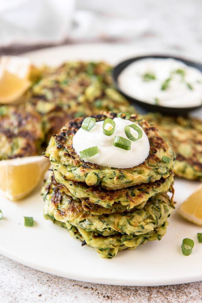 a stack of zucchini fritters topped with a dollop of sour cream and sliced green onions. More fritters, a dish of sour cream and lemon wedges in the background.