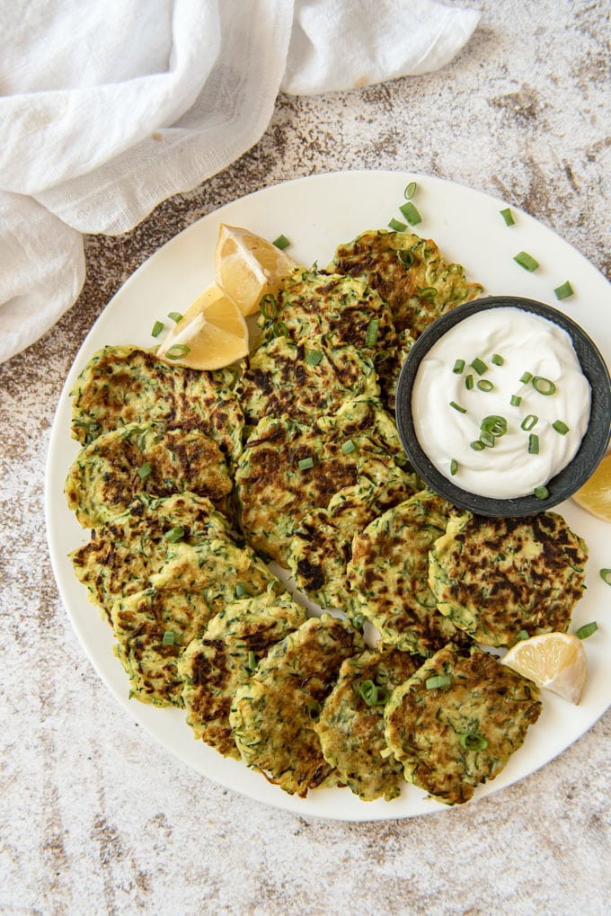 An overhead shot of zucchini fritters on a white plate with lemon wedges and a dish of sour cream topped with sliced green onions.