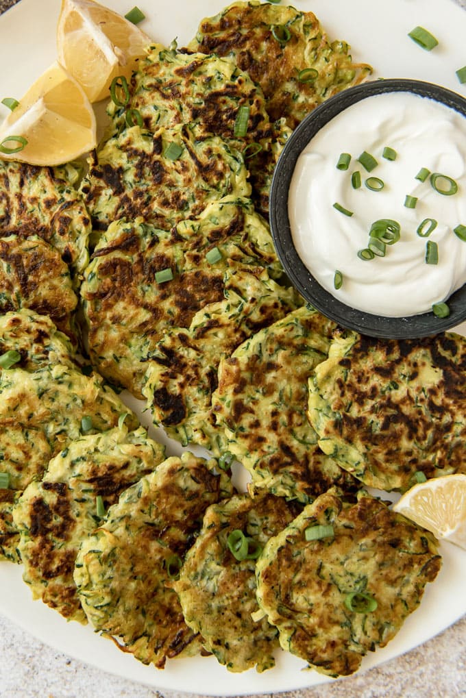 An overhead of zucchini fritters on a white plate wit a dish of sour cream and lemon wedges.