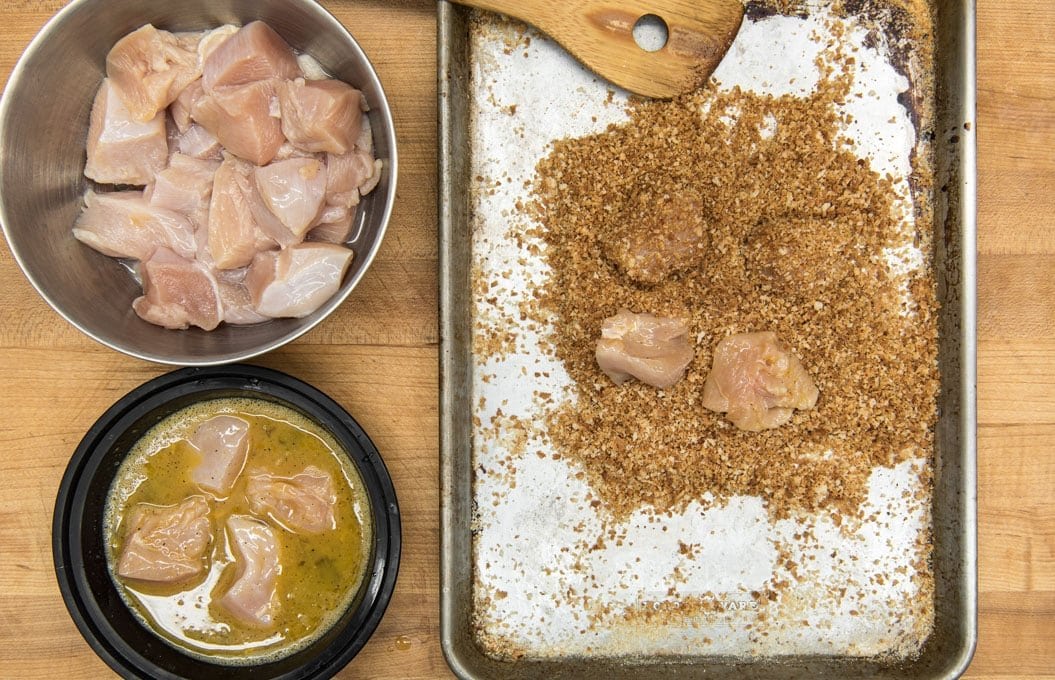 a wood cutting board background, a silver bowl with raw chicken nuggets, a black dish with whisked egg and 4 pieces of chicken and a silver baking sheet with a wood spoon, breadcrumbs and 3 pieces of raw chicken.