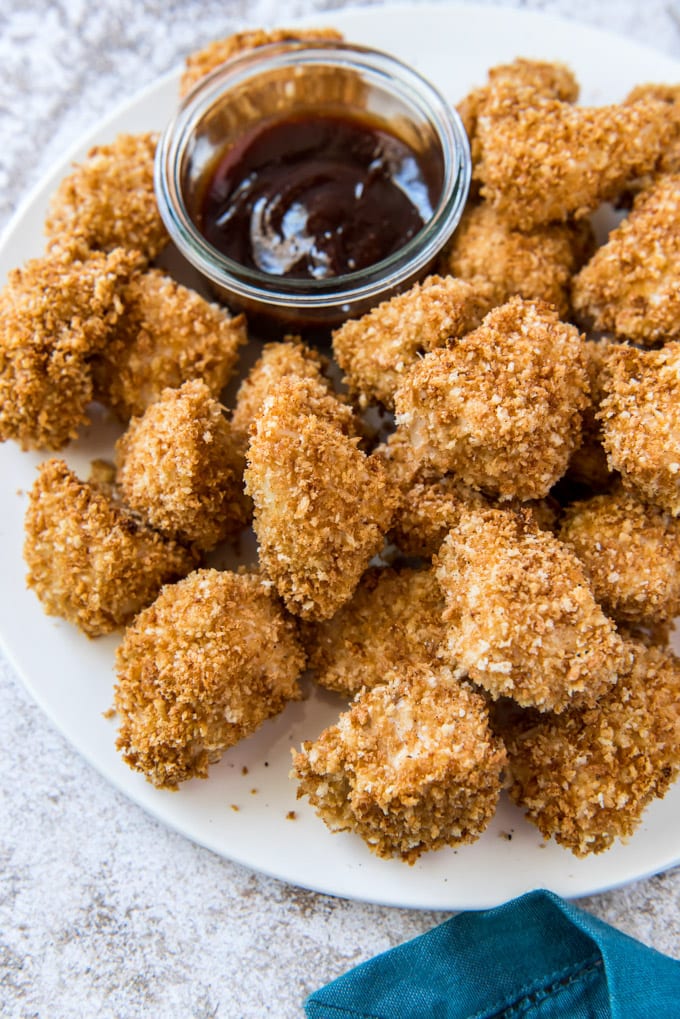 a white plate full of baked chicken nuggets, a clear glass dish with barbecue sauce and a blue napkin on a gray background
