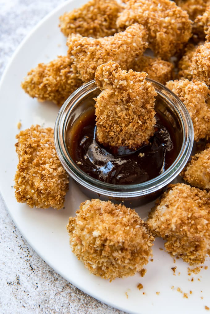 a white platter with a bunch of chicken nuggets and a dish of bbq sauce on a gray and white background