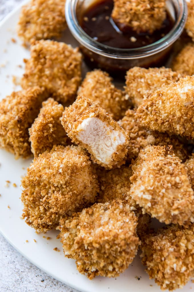 a white plate with a pile of homemade chicken nuggets and a dish of bbq sauce. one nugget has a bite taken out of it.