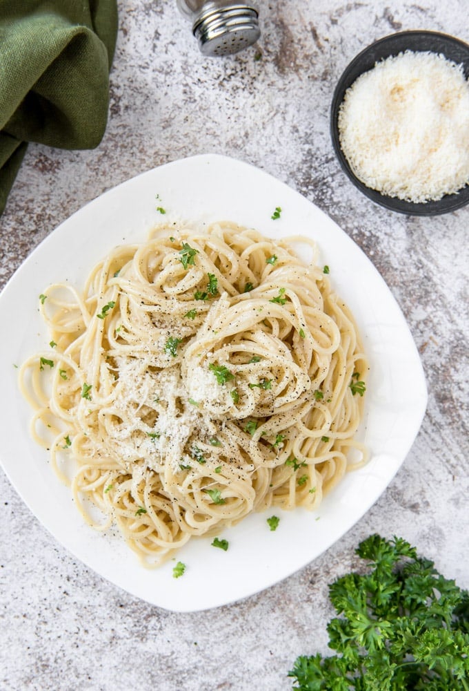 An overhead image of a white plate with cacio e pepe (spaghetti), a small bowl of parmesan cheese, and parsley on a gray background.
