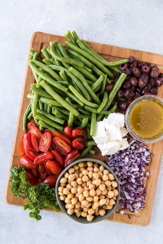 A wooden cutting board with green beans, olives, tomatoes, chickpeas, parsley, onions, feta cheese and a bowl of dressing. 