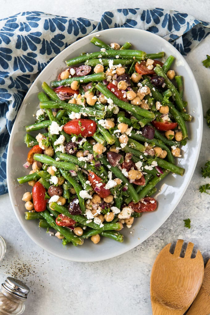 An overhead image of a white plate filled with green bean, tomatoes, olives, chickpeas and feta cheese. A blue and white napkin, a wooden spoon and salt and pepper shakes sit off to the sides.