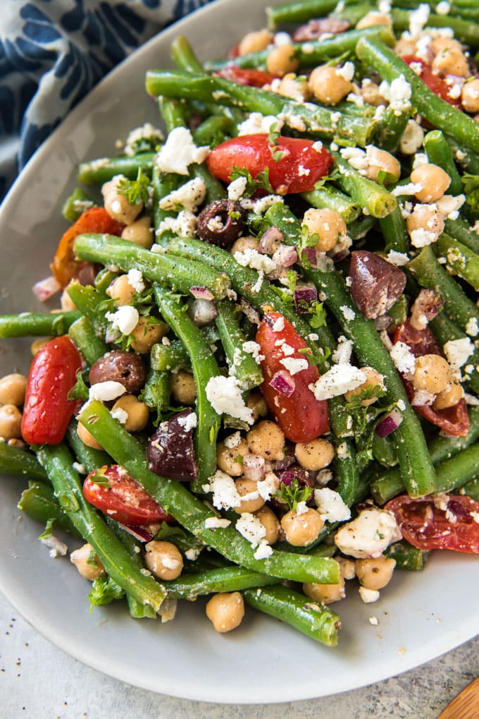 A close up overhead image of a white plate filled with green bean, tomatoes, olives, chickpeas and feta cheese. A blue and white napkin sits off to the sides.