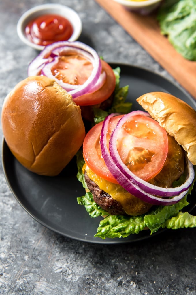 Two hamburgers on a plate, topped with cheese, tomatoes, onions and lettuce. The tops of the buns are angled off the edge of the burgers.