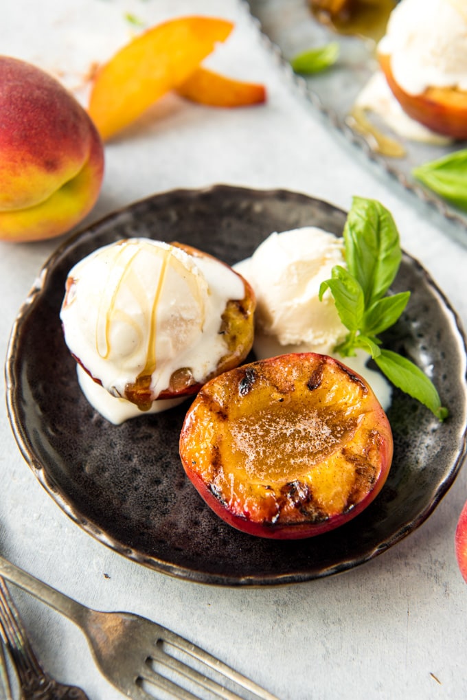 2 peaches halves on a black plate, one has vanilla ice cream and a honey drizzle. Mint leave, a whole peach and a few peach slices are also pictured.
