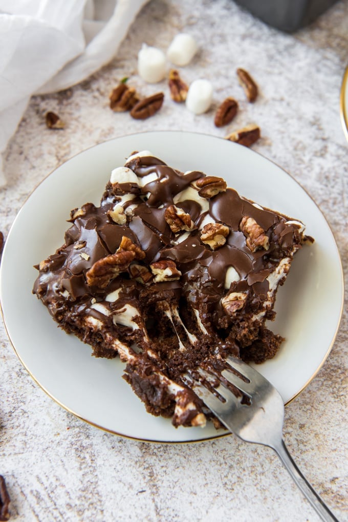 a slice of Mississippi mud cake on a white plate with a fork digging in. Marshmallows and pecans are scattered around on a grey and white background.