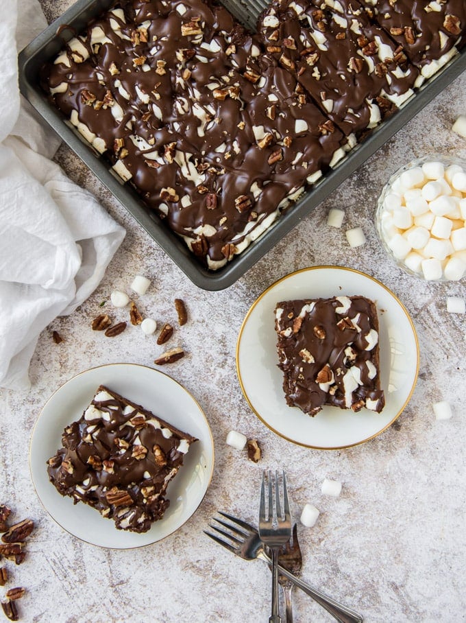 an overhead image of Mississippi Mud Cake in a large pan, with two square pieces of the cake on white plate. Scattered around on the gray background are marshmallows, pecans and a few forks.