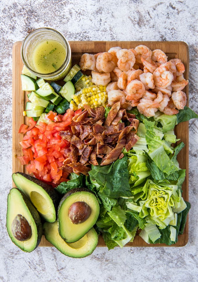 a wood cutting board, 4 avocado halves, chopped lettuce, diced tomatoes, chopped cucumber, shrimp, bacon and corn. a clear glass jar with yellow dressing.