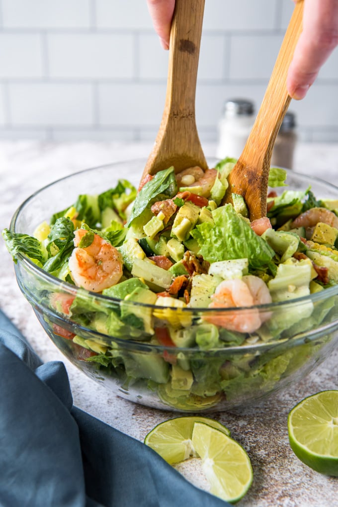 a glass bowl, salad with shrimp, bacon and corn, limes slices, a blue napkins, hands holding wooden spoons in the salad.