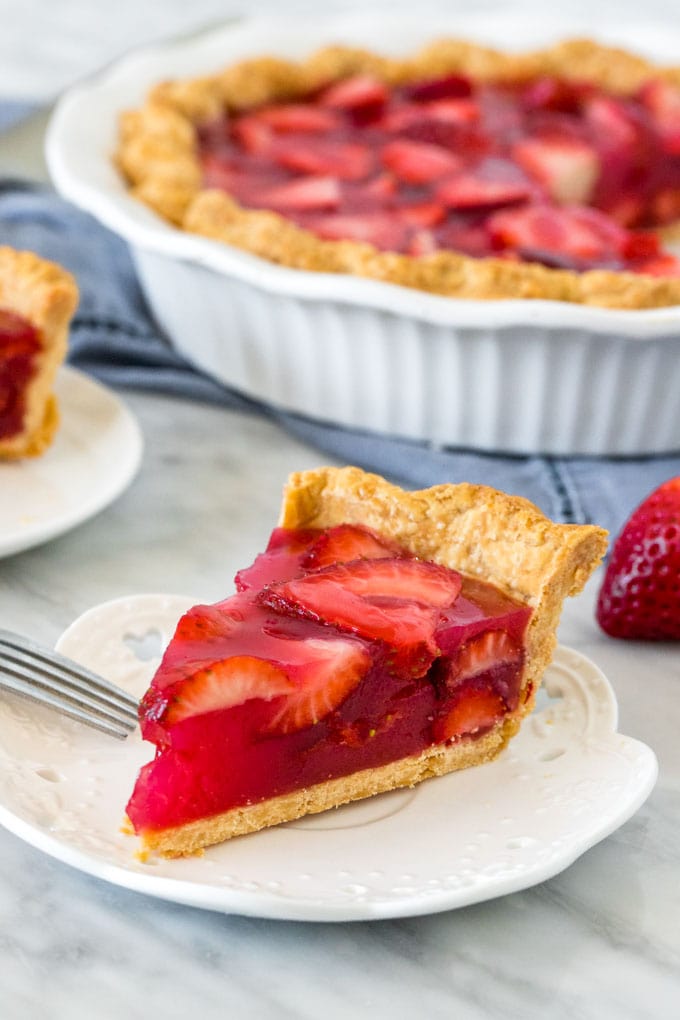 Slice of strawberry jello pie on a white plate. a whole strawberry pie in a white dish. A blue napkin.